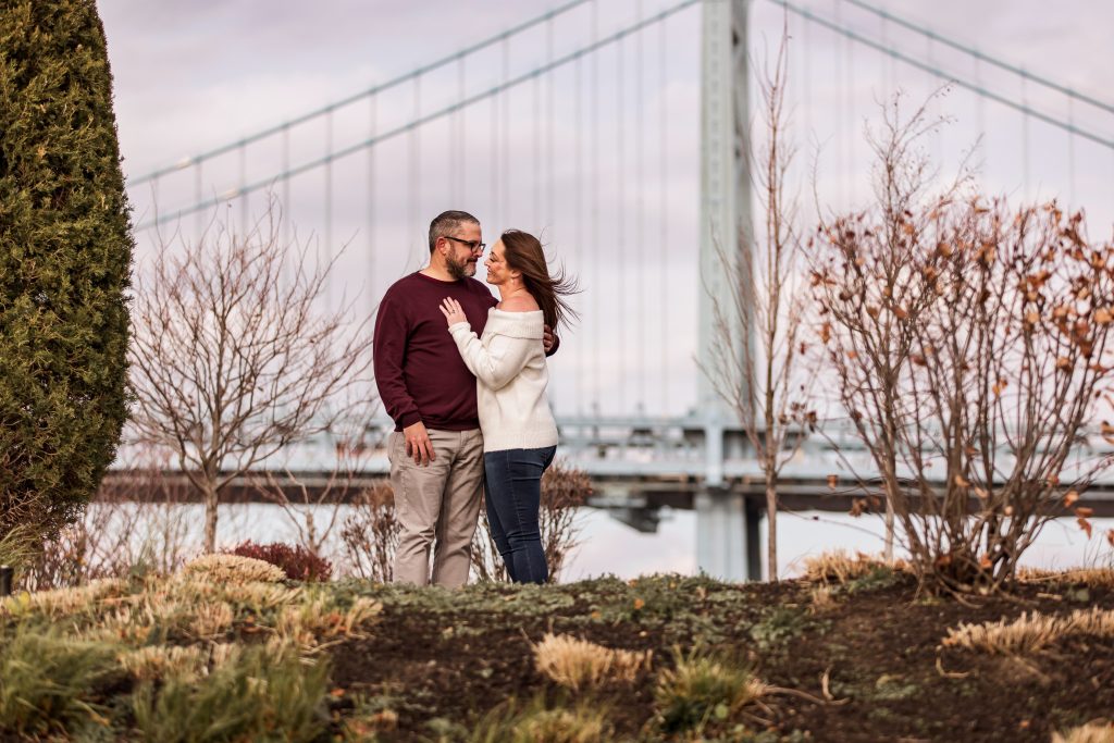 Engaged couple posing together in front of the Philadelphia bridge during their engagement photo shoot.